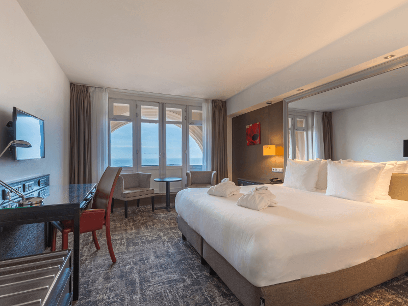 Superior kamer in Grand Hotel Amrâth Kurhaus met twee eenpersoonsbedden met twee badjassen. In de hoek van de kamer staan twee grijze stoelen met een zwart tafeltje daarnaast een zwart bureau met een rode stoel