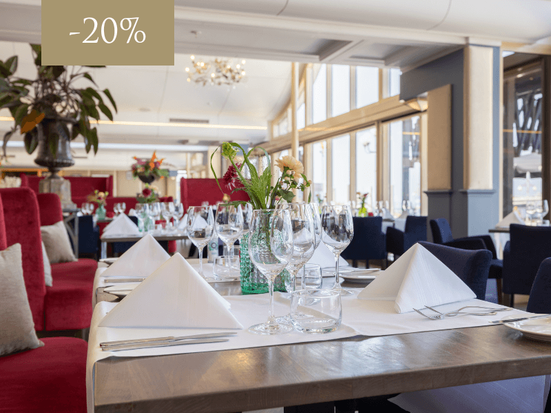 Restaurant Waves at the Kurhaus, with large windows and a view of the water. Elegant tables with white linen and flowers.