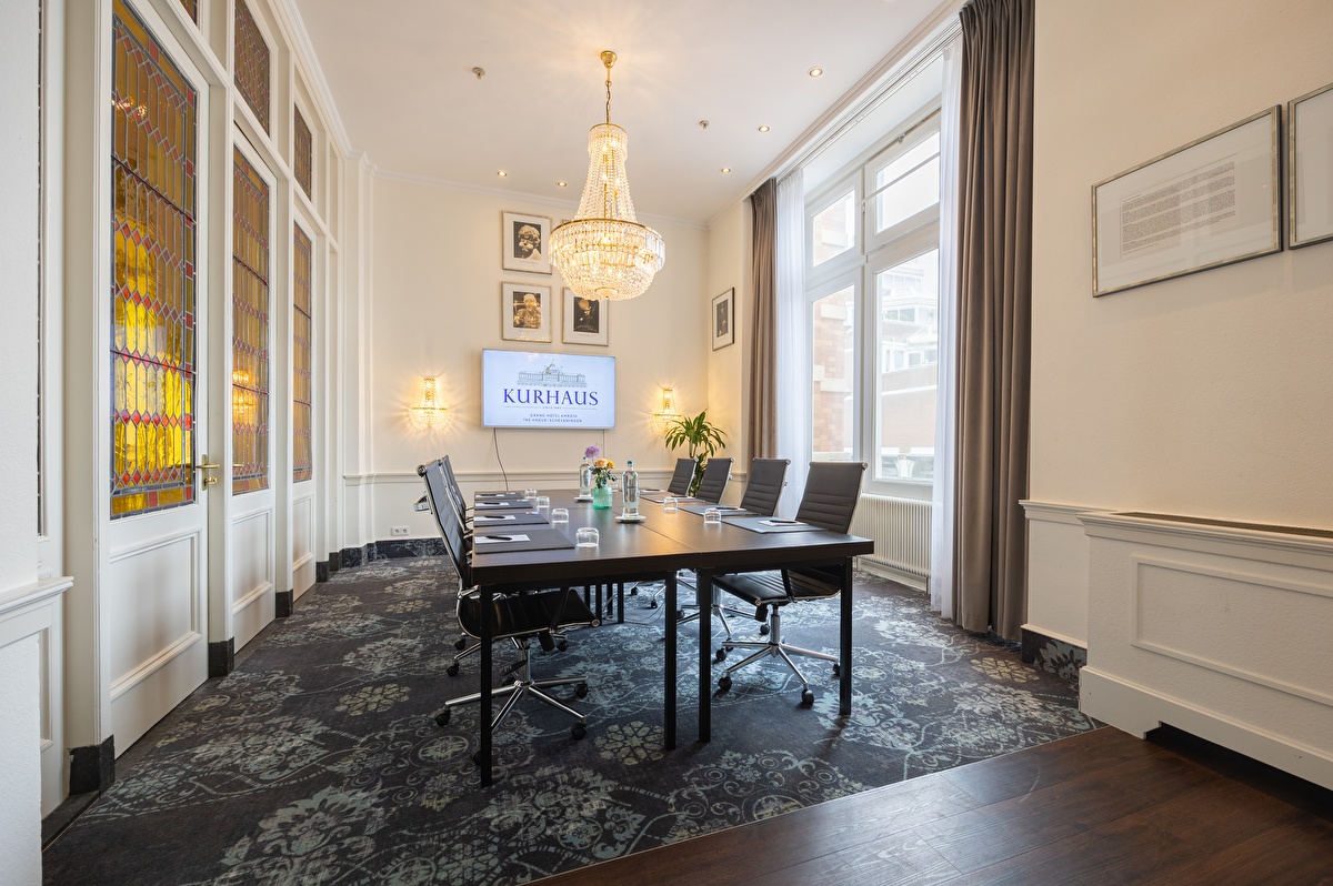 Paul van Vliet meeting room with a table, leather chairs, crystal chandelier, and a screen with the Kurhaus logo. Large windows and stained-glass doors provide plenty of natural light.