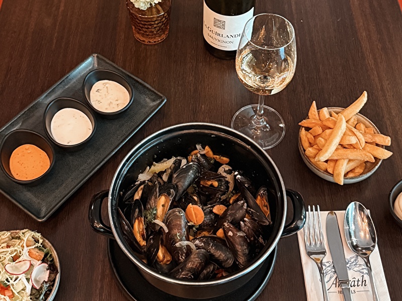 Geserveerde pan met verse mosselen in een zwarte pan, drie sausjes, friet, salade en een glas witte wijn (La Guirlande Sauvignon Blanc) op een donkerhouten tafel bij Amrâth Hotels.