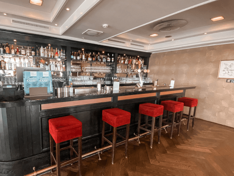 Hotel bar in Grand Hotel Amrâth Kurhaus with red bar stools, a dark wooden bar, and a well-stocked liquor cabinet.