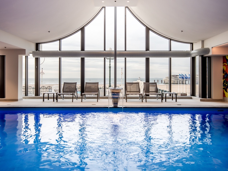 Wellnessruimte met een binnenzwembad, ligstoelen en grote ramen met uitzicht op de zee. De inrichting bevat planten en een wanddecoratie met een historisch tafereel.