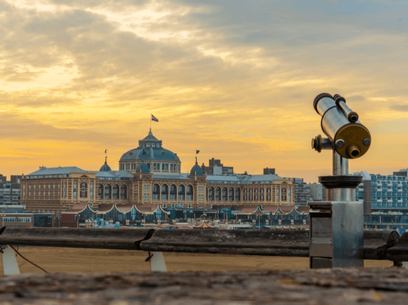 Uitzicht op Grand Hotel Amrâth Kurhaus in Scheveningen bij zonsondergang, met een telescoop op de voorgrond en een kleurrijke lucht op de achtergrond.